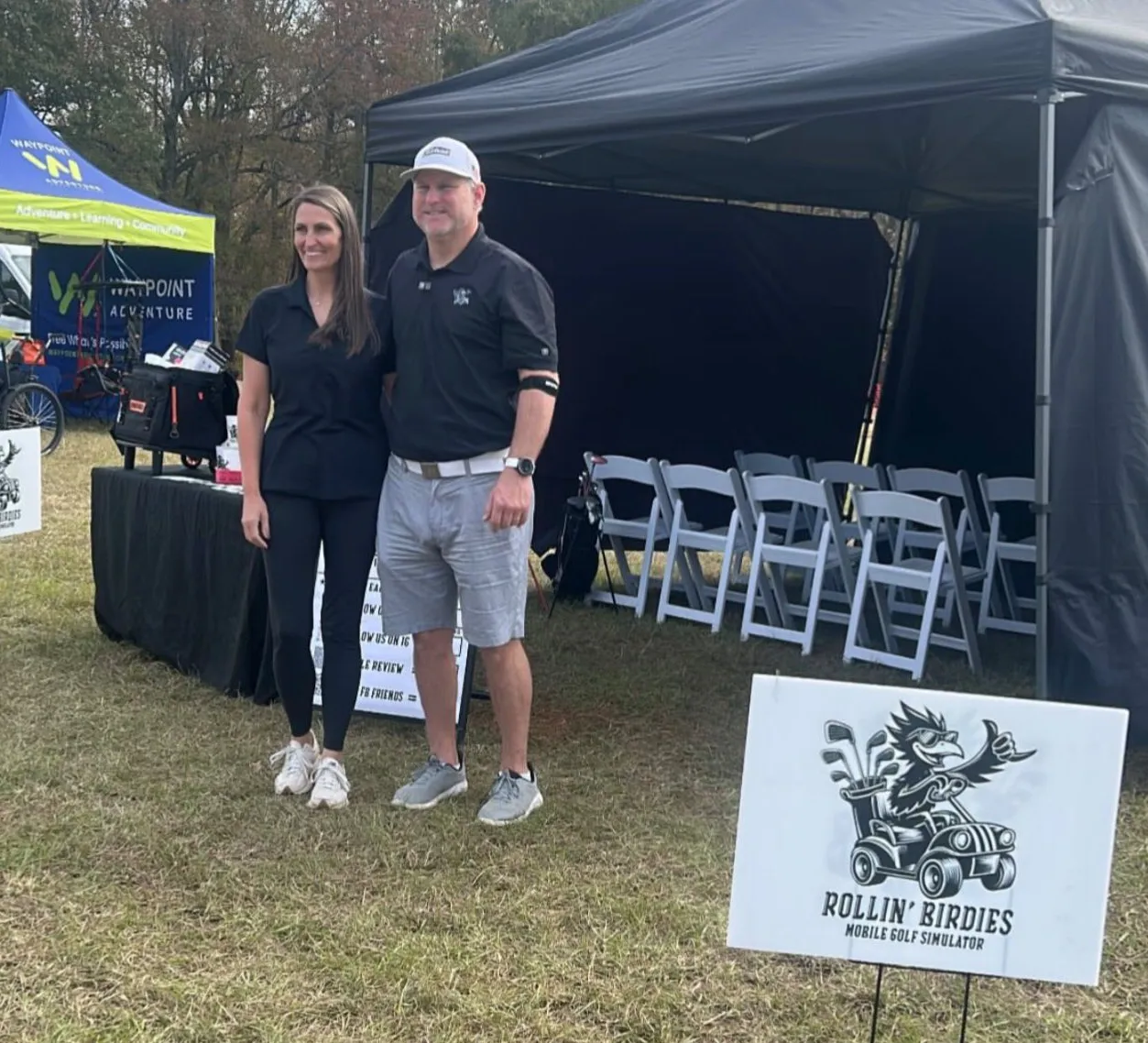 about-1 A man and woman stand smiling outside a black tent at an outdoor event with golf-themed signage.