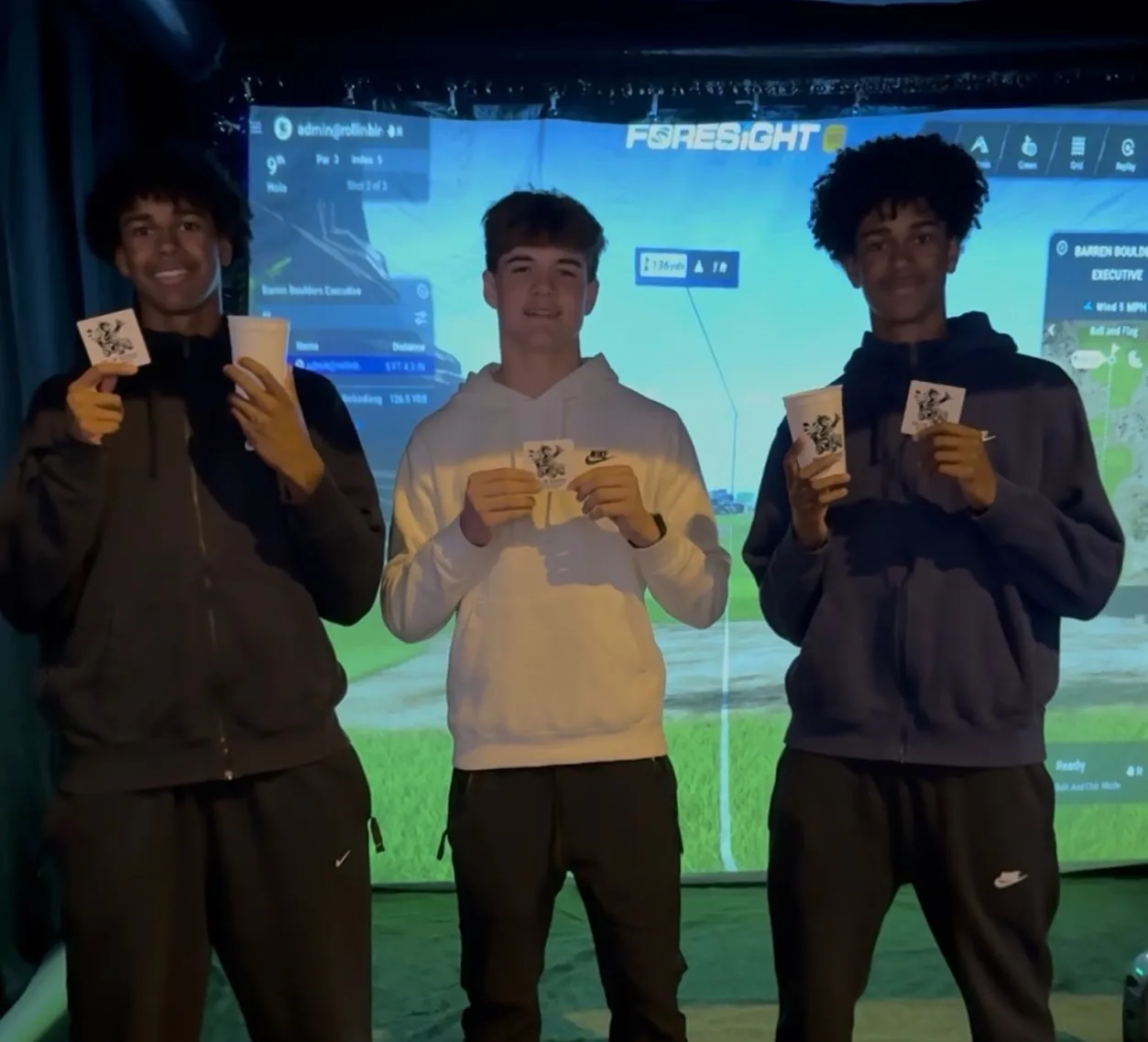 about-2 Three young men stand indoors holding cups and cards, smiling, with a golf simulator screen in the background.