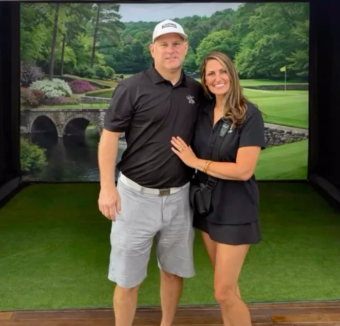 about-3 A smiling couple poses in golf attire on a green with a scenic golf course backdrop.