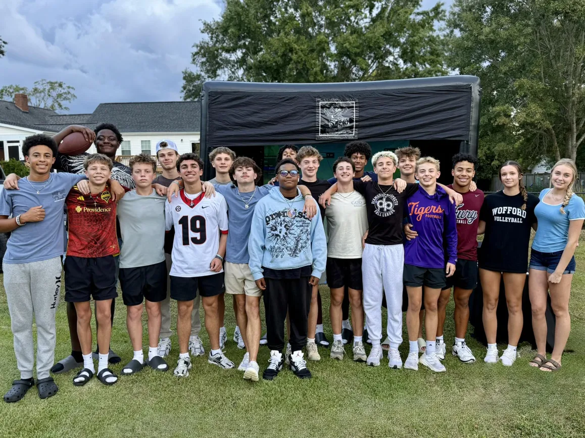 A group of teens poses together outside in front of a large inflatable screen on a grassy lawn.