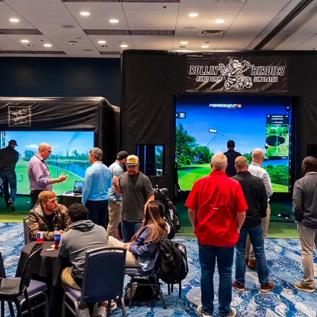 People gather around golf simulators at an indoor event, with some playing and others watching or talking.