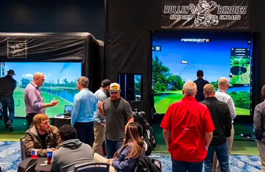 People gathered around indoor golf simulators, some playing while others watch and converse.