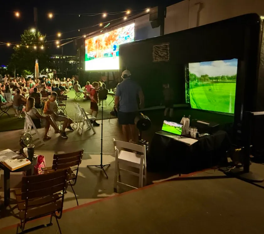 event-1 People gather outdoors at night with string lights, tables, and a golf simulator setup in the foreground.