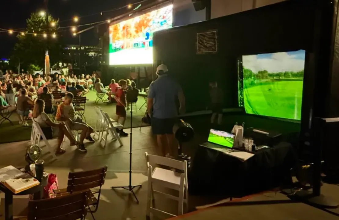 People play golf on a simulator at an outdoor event with string lights and a seated audience at night.