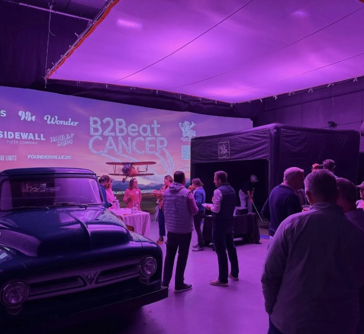 People gather at an indoor B2Beat Cancer event with purple lighting and a vintage truck on display.