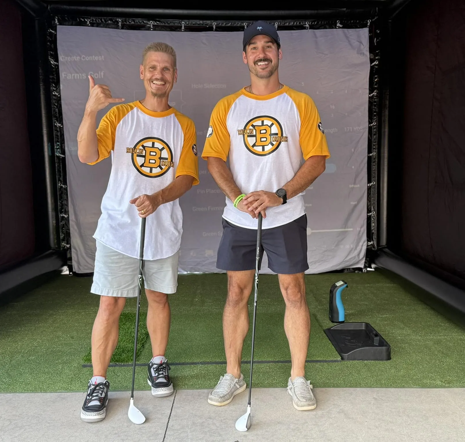 festival-2 Two men in matching baseball shirts and holding golf clubs stand and smile in an indoor golf simulation area.