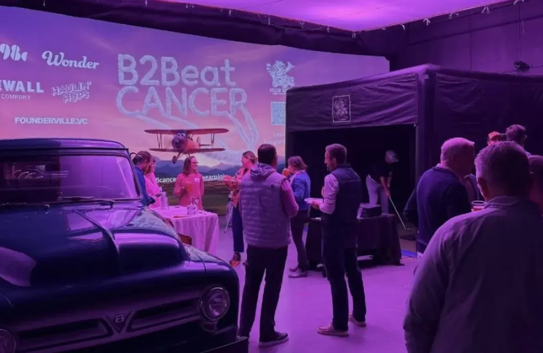 People gather at an indoor event called "B2Beat Cancer," with purple lighting and a vintage car on display.