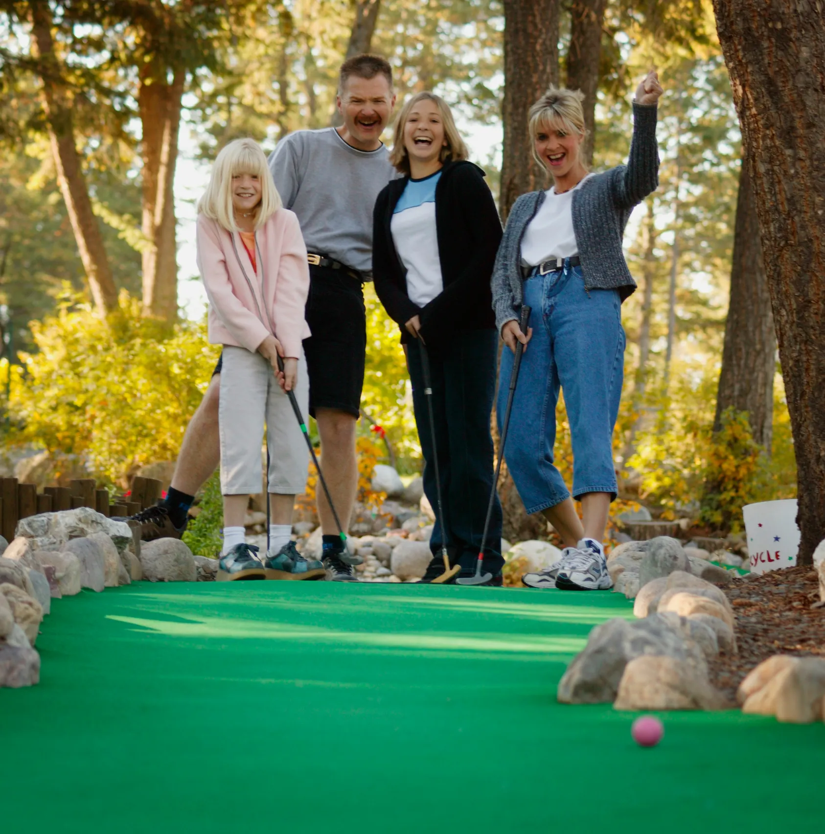 Four people smiling and playing mini golf on a green course surrounded by rocks and trees.