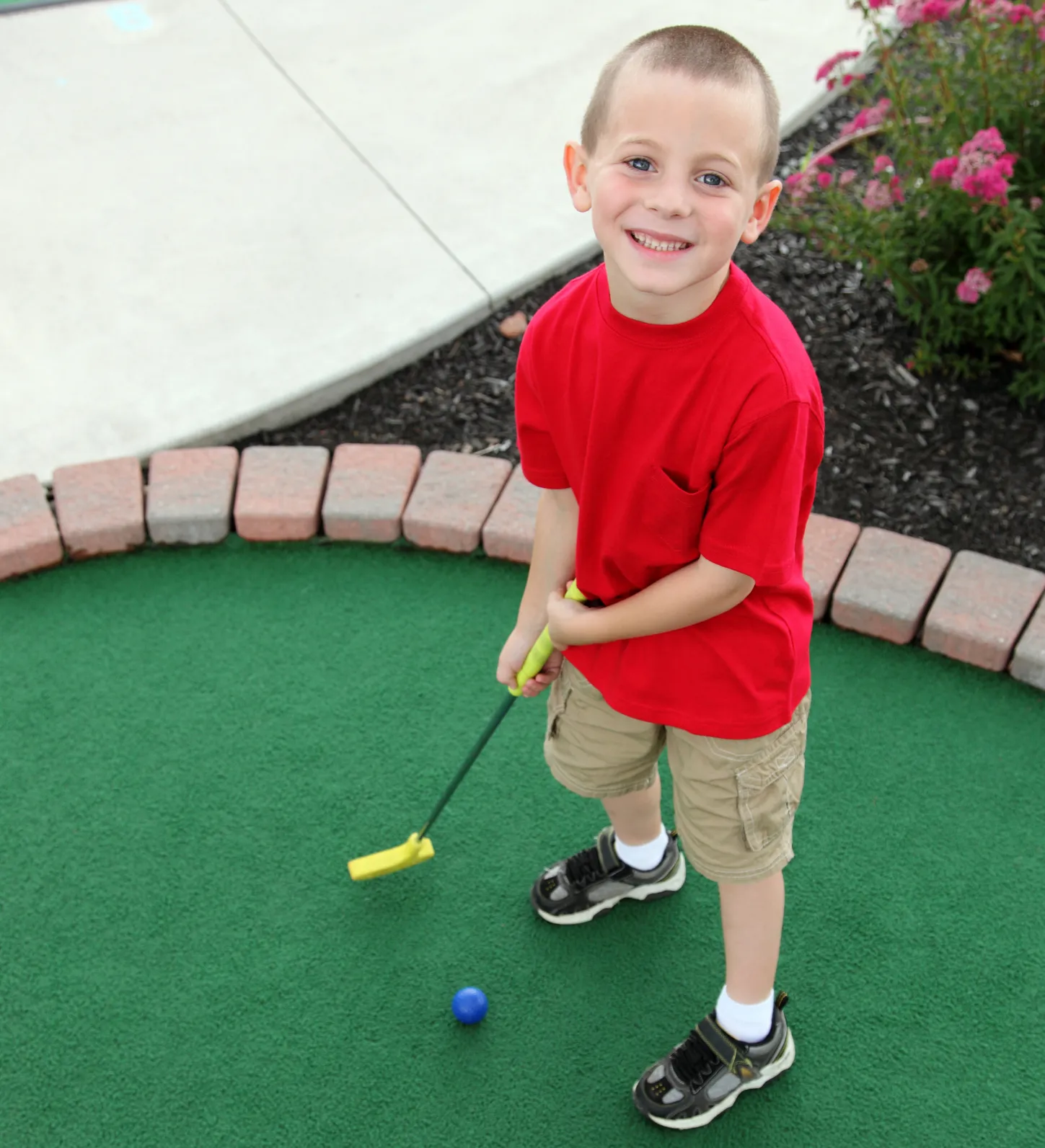 Smiling boy in a red shirt plays mini golf, holding a yellow putter near a blue golf ball on green turf.