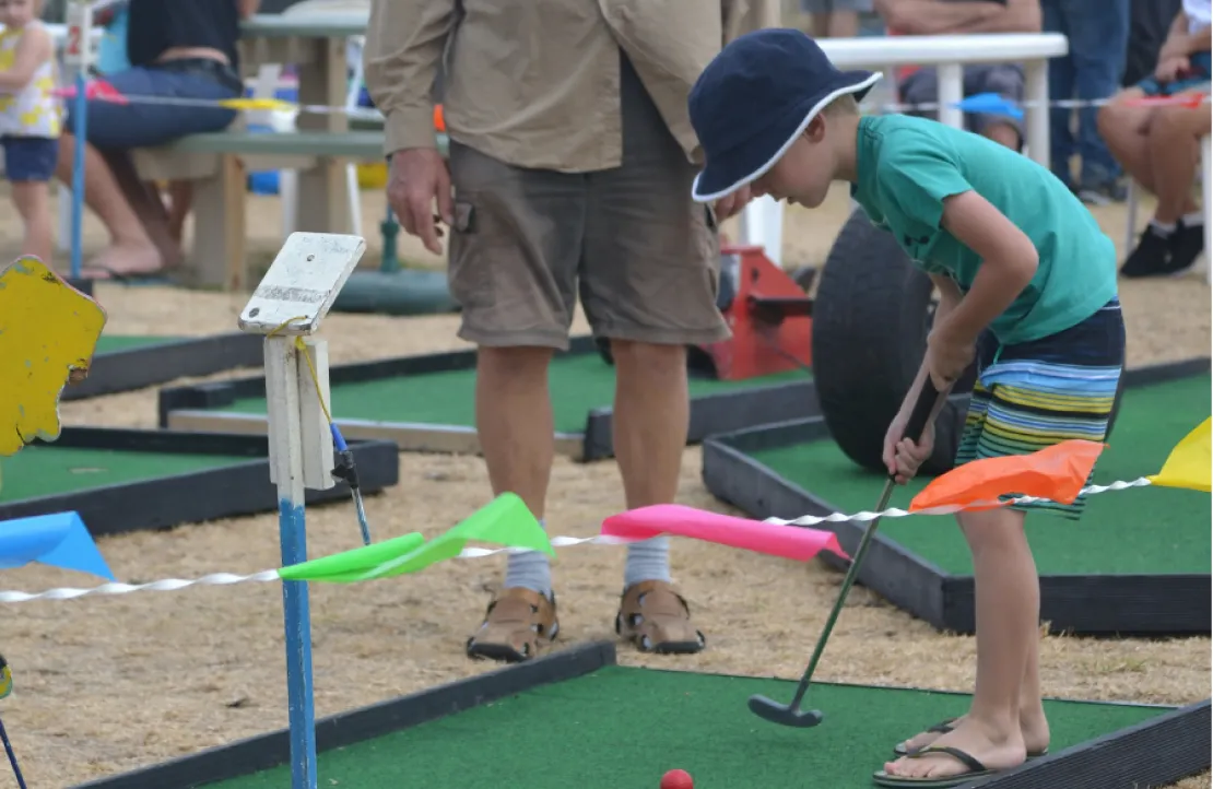 A young boy in a hat plays mini-golf, aiming his putter at a red ball on a green course.