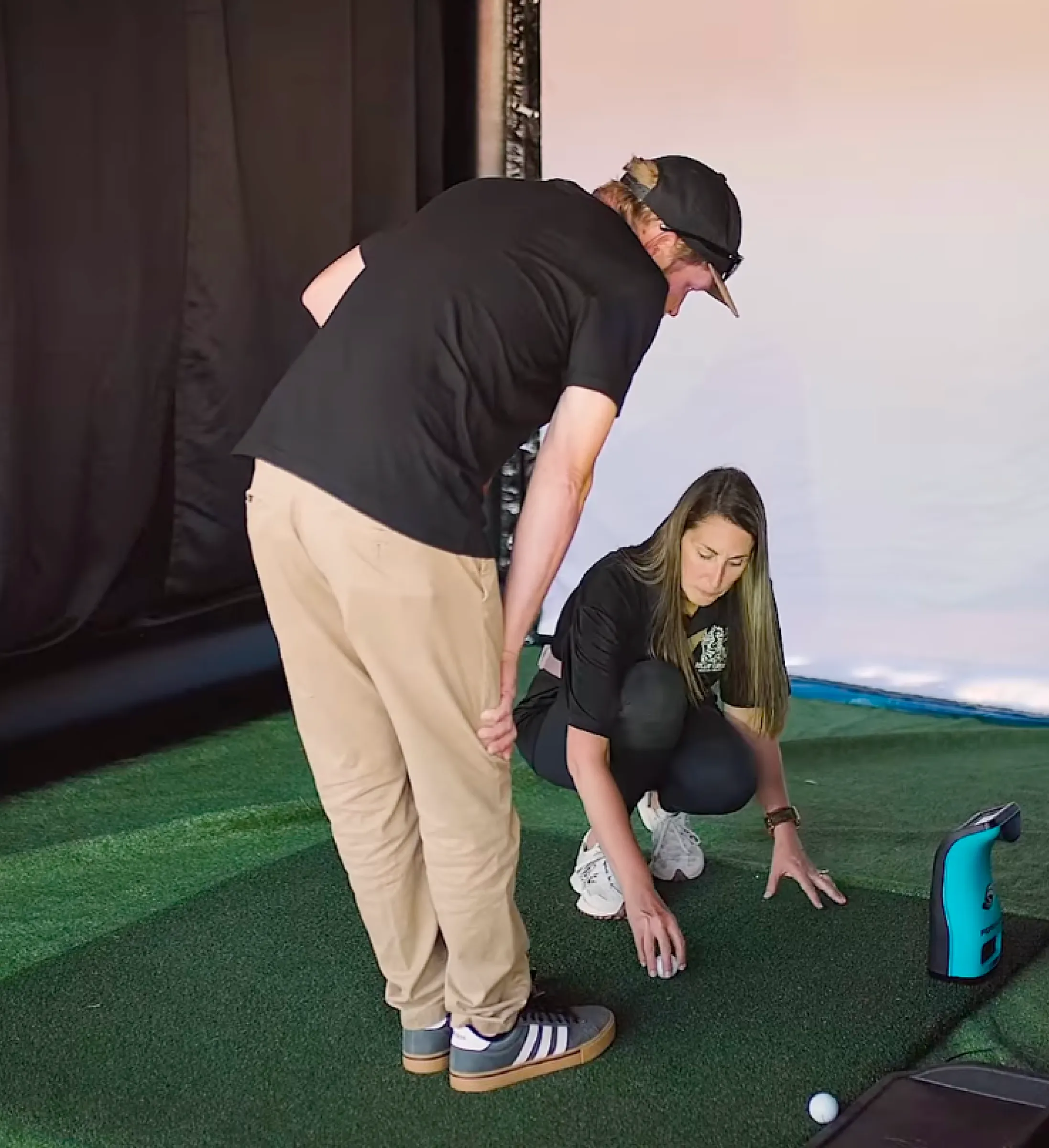 A woman crouches to place a golf ball on artificial turf as a man watches beside her indoors.