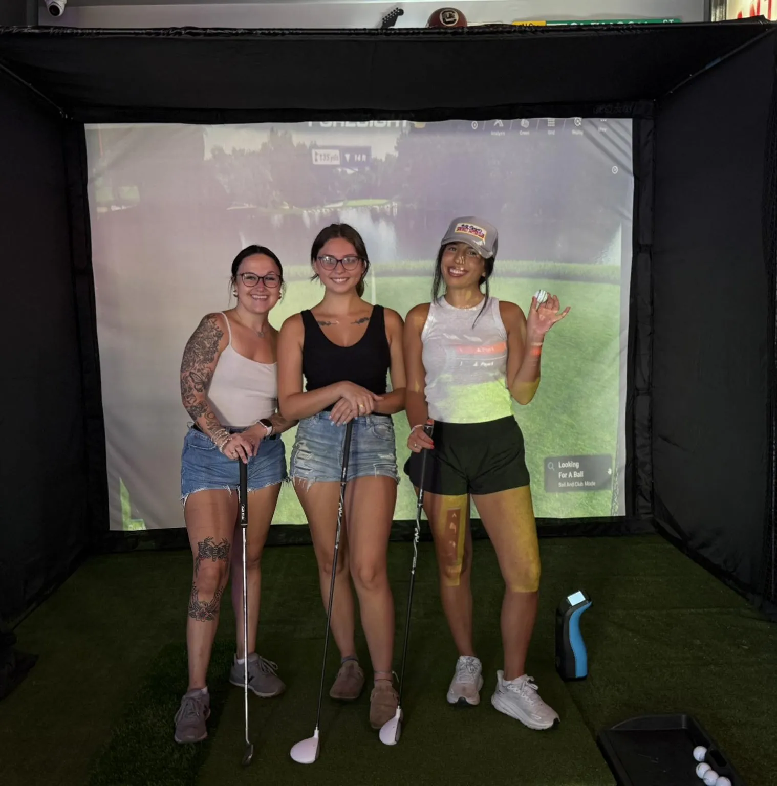 Three women pose with golf clubs in front of an indoor golf simulator screen, smiling at the camera.