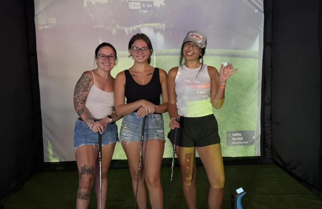 Three women in casual summer outfits pose with golf clubs in front of a virtual golf screen, smiling at the camera.