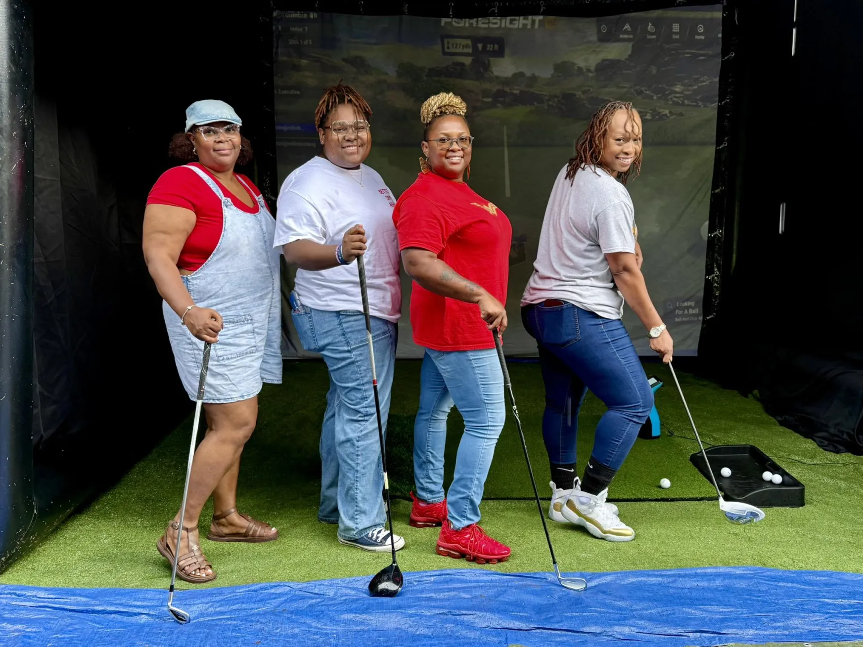 Four women pose with golf clubs on an indoor green, smiling in front of a golf simulator screen.