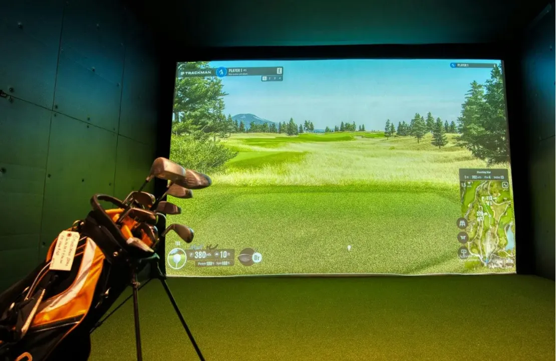 Golf simulator with virtual golf course on a screen and a golf bag with clubs in the foreground.