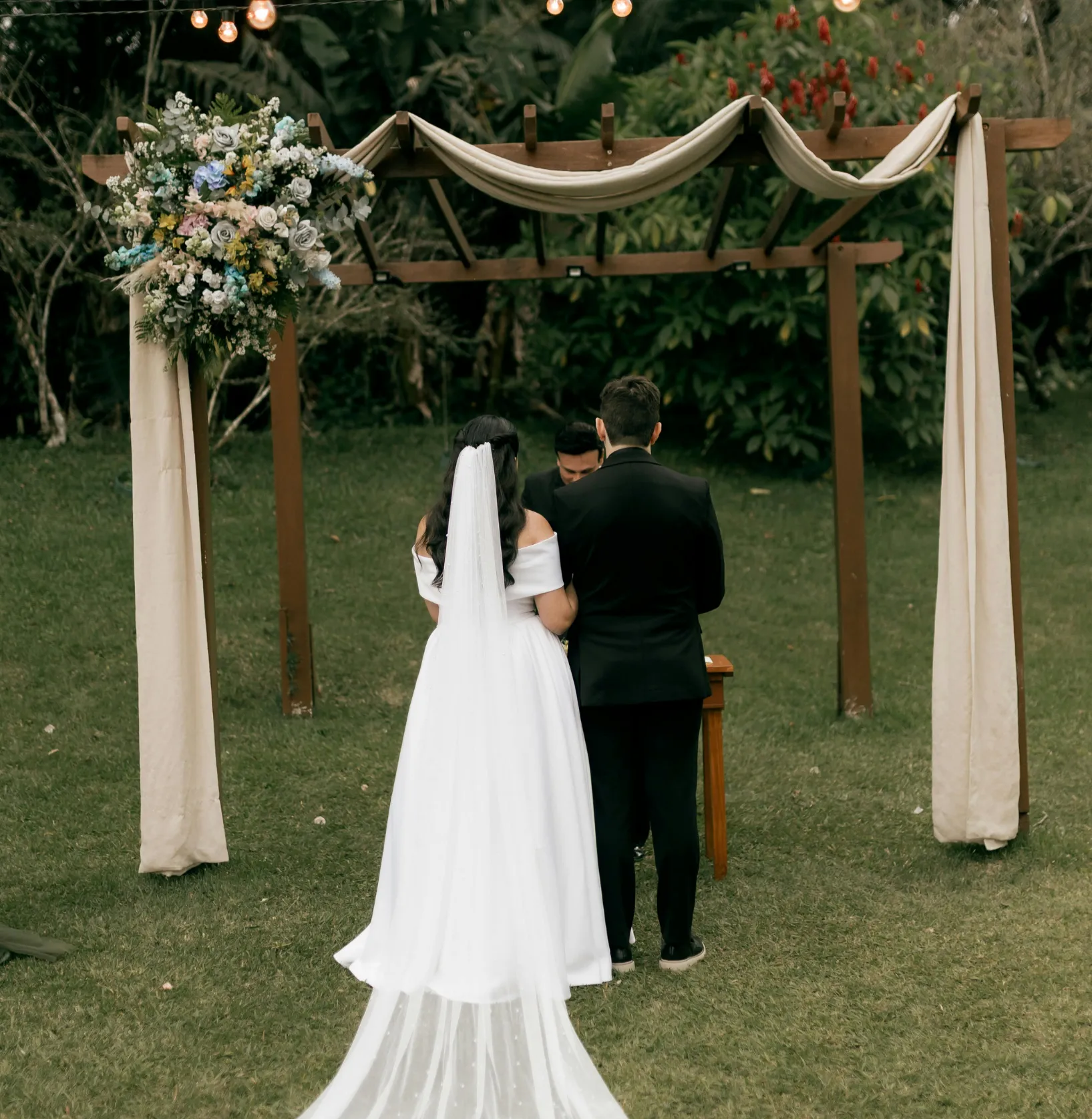 wedding-1 Bride and groom stand together at an outdoor altar decorated with flowers and draped fabric, seen from behind.