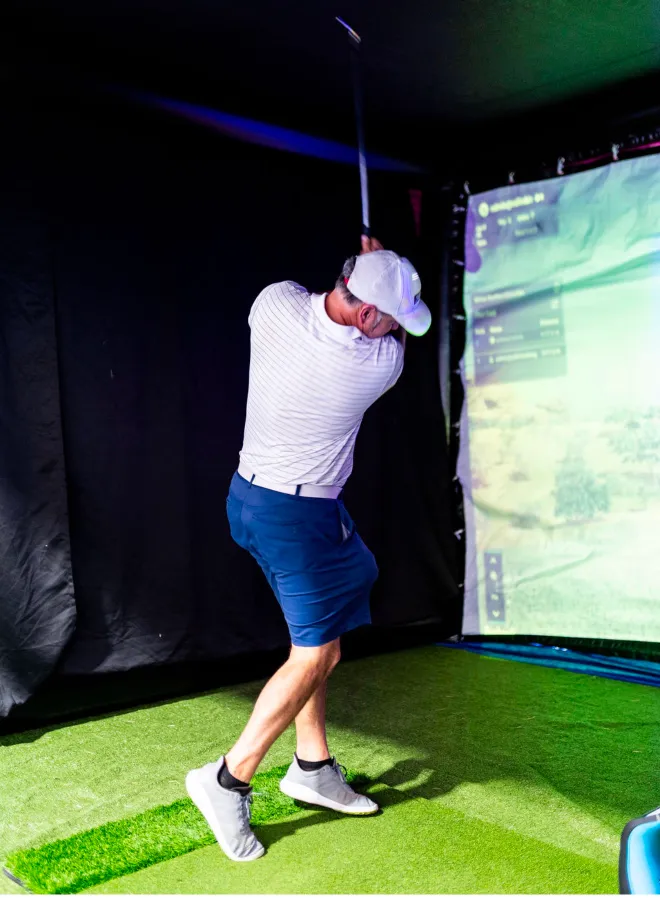 why-choose-1 Man swinging a golf club indoors on artificial grass, aiming at a virtual golf simulator screen.